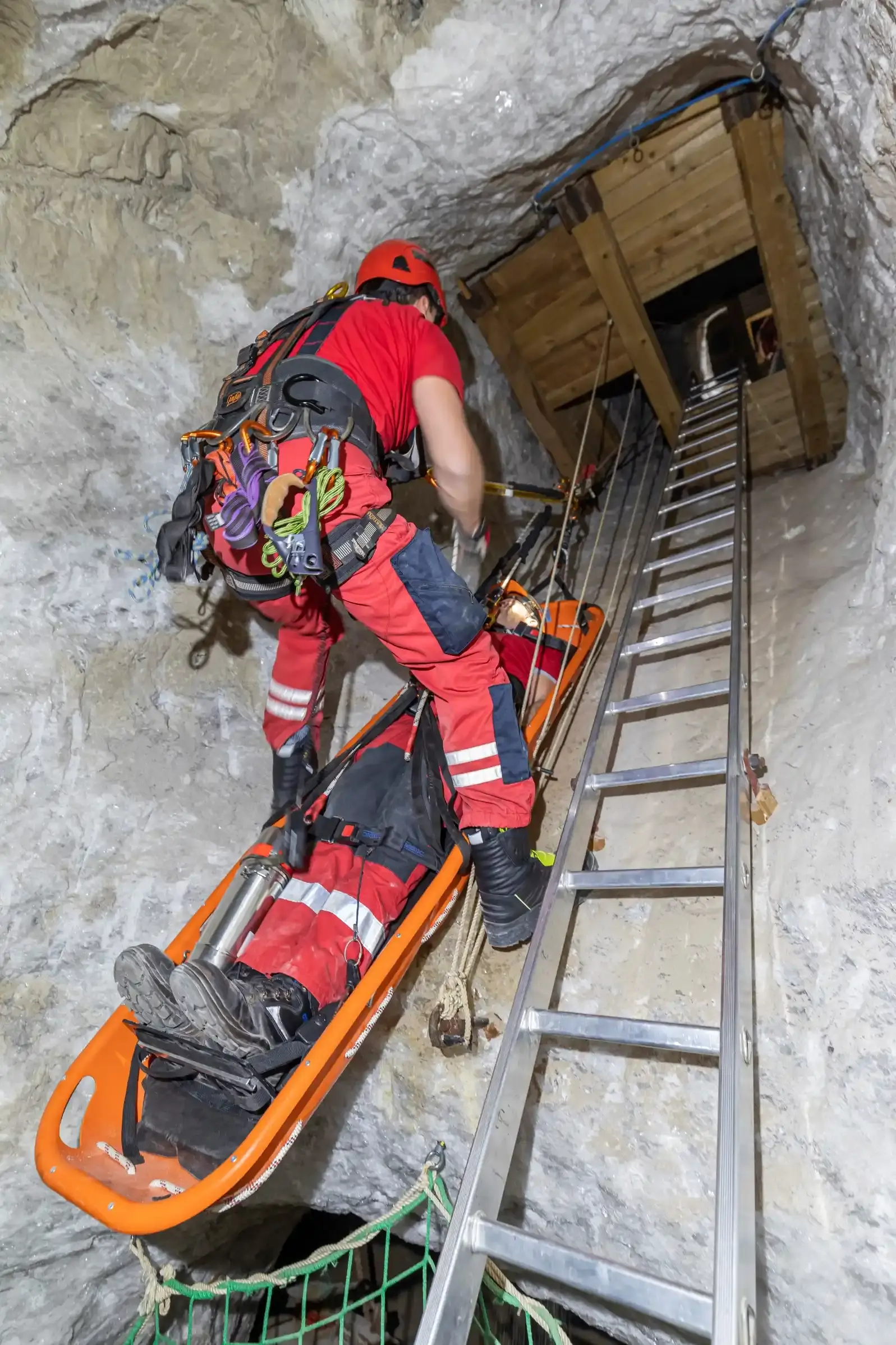 In einem senkrecht verlaufenden Schacht eines Bergwerks befördert eine an Seilen befestigte Person eine andere Person auf einer Bahre. Neben ihnen verläuft eine lange Leiter. Es handelt sich um eine Übung der Höhenrettung. 