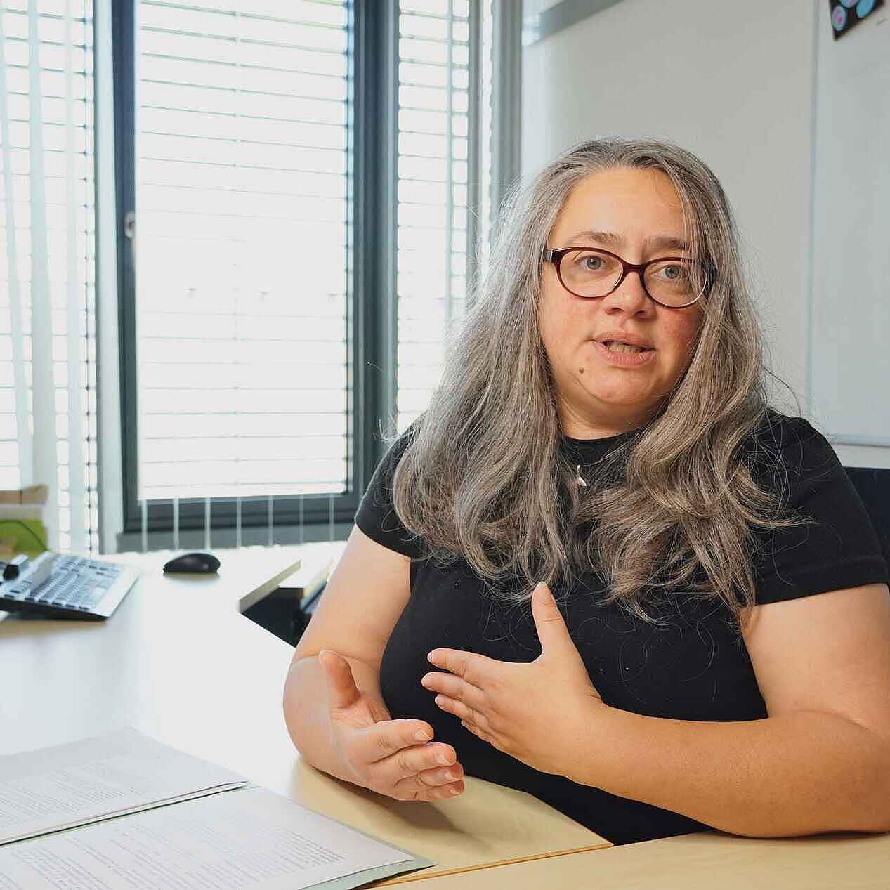 Eine Frau mit langen grauen Haaren und Brille sitzt hinter einem Schreibtisch. Ihre Hände gestikulieren. 