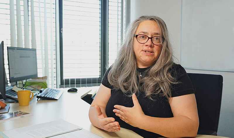 Eine Frau mit langen grauen Haaren und Brille sitzt hinter einem Schreibtisch. Ihre Hände gestikulieren. 
