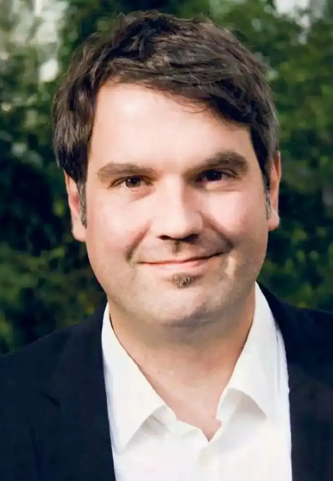 Portraitfoto eines Mannes mit dunkelbraunen Haaren, weißem Hemd und schwarzem Sakko.