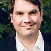 Portraitfoto eines Mannes mit dunkelbraunen Haaren, weißem Hemd und schwarzem Sakko.