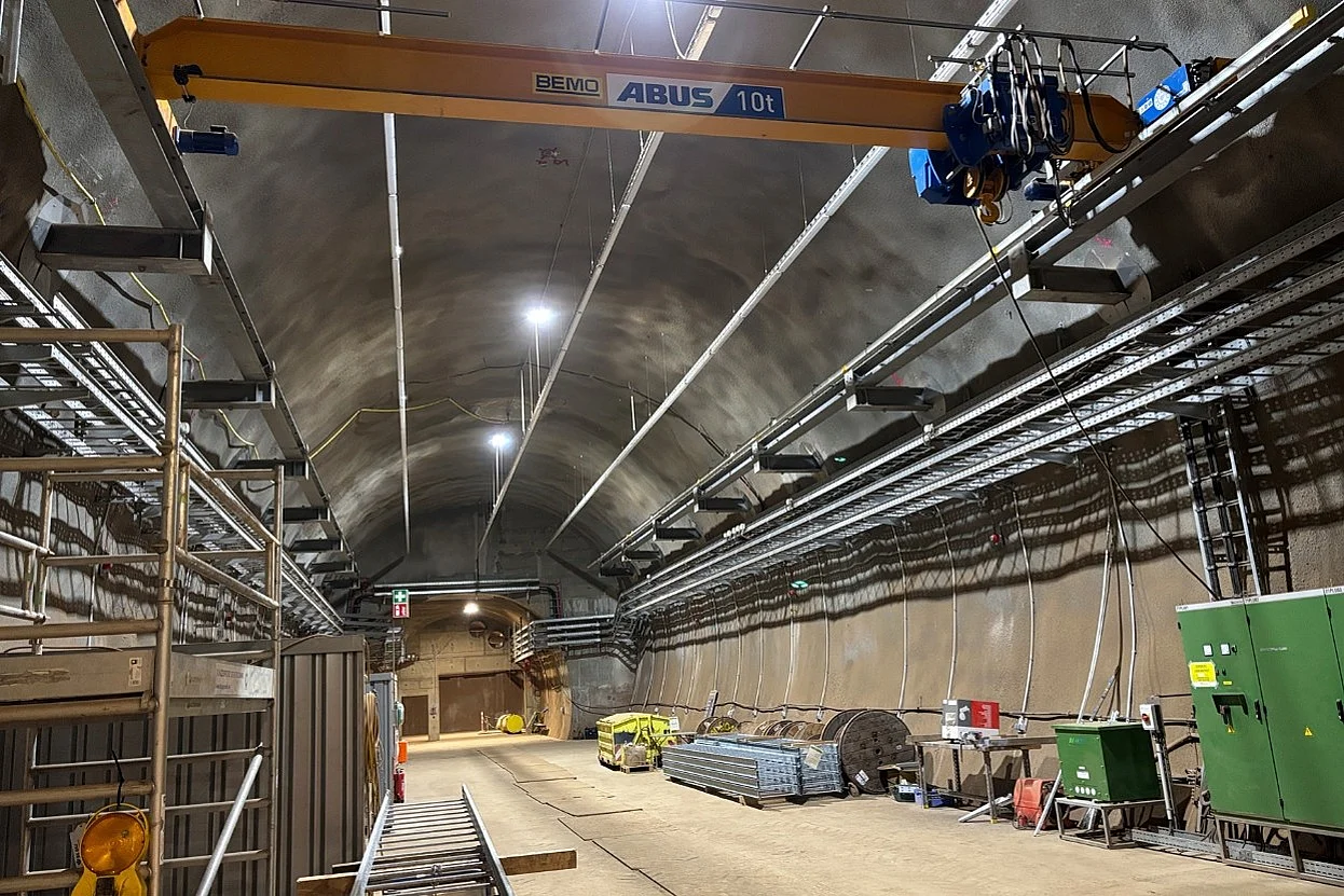 Blick in einen Tunnel in einem Bergwerk. An den Wänden verlaufen Rohre und Gitterkonstruktionen sowie Schienen mit einer gelben Kranbrücke.