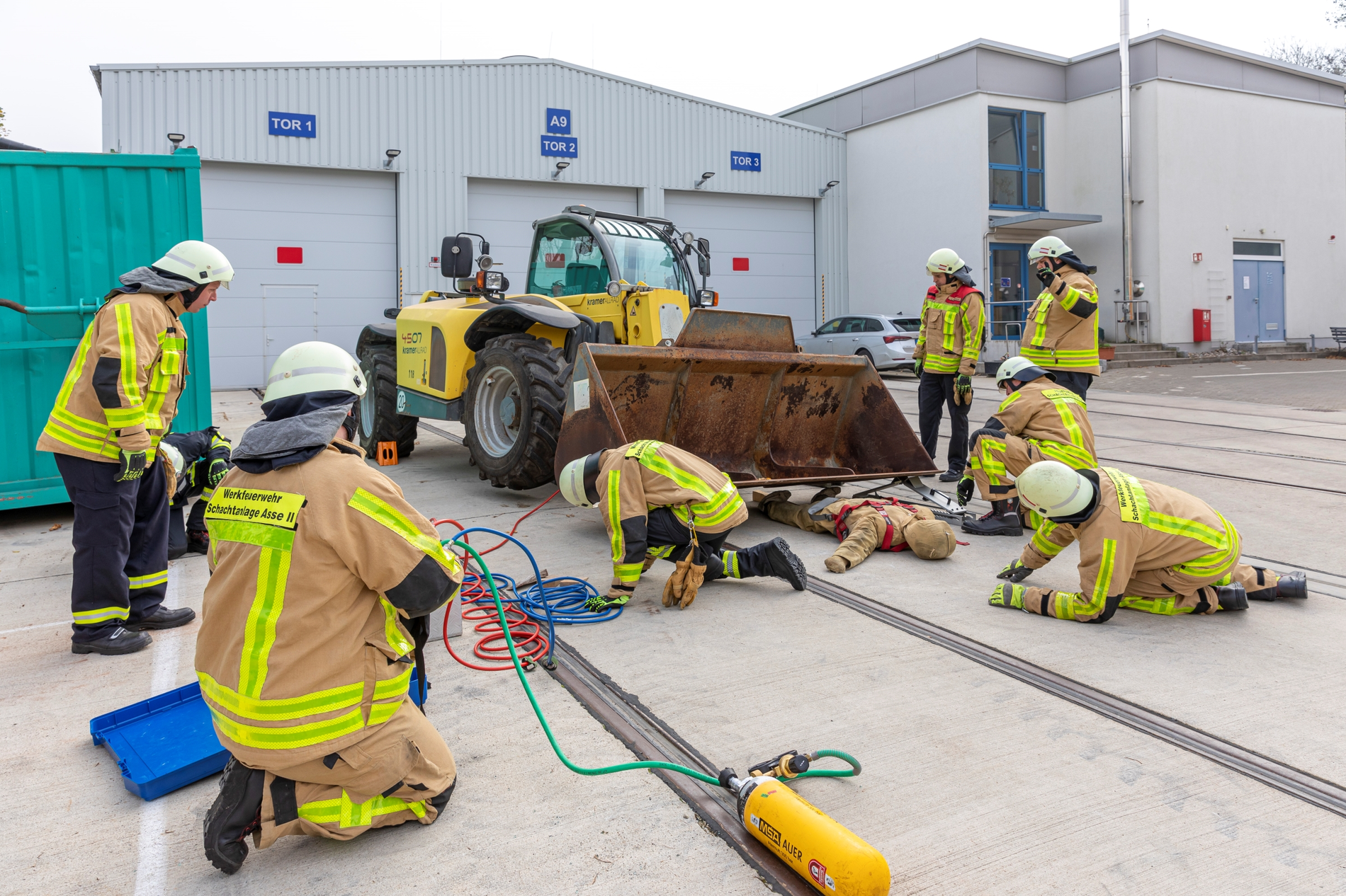 Vor einem großen Radlader findet eine Übung der Werksfeuerwehr statt. Mehrere Mitglieder der Wehr stehen und knien um eine scheinbar verletzte Person herum, die unter der Radladerschafel liegt. Bei der verletzten Person handelt es sich um eine Puppe.