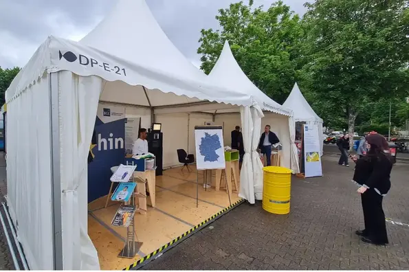 Ein weißer Pavillon mit Holzboden, Schautafeln und Magazinständern. Vor dem Pavillon steht ein gelbes Fass und eine Besucherin in schwarzer Kleidung.