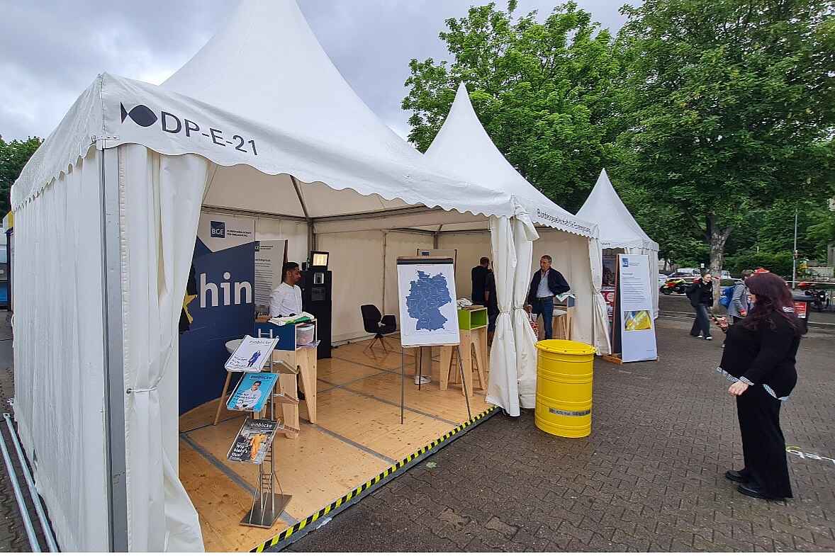Ein weißer Pavillon mit Holzboden, Schautafeln und Magazinständern. Vor dem Pavillon steht ein gelbes Fass und eine Besucherin in schwarzer Kleidung.