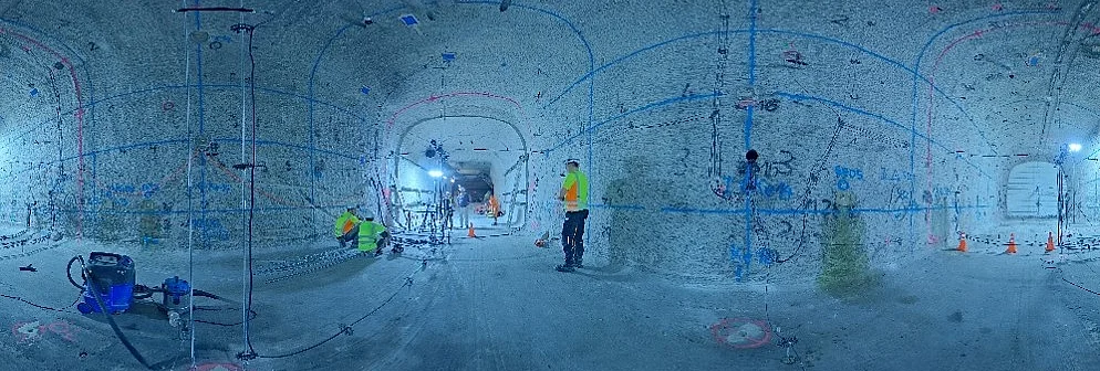 Ein untertägiger Hohlraum ist an den Wänden mit blauen und roten horizontalen und vertikalen Linien versehen. Mehrere Personen mit Warnwesten und mit Schutzhelmen bedienen verschiedene technische Geräte.