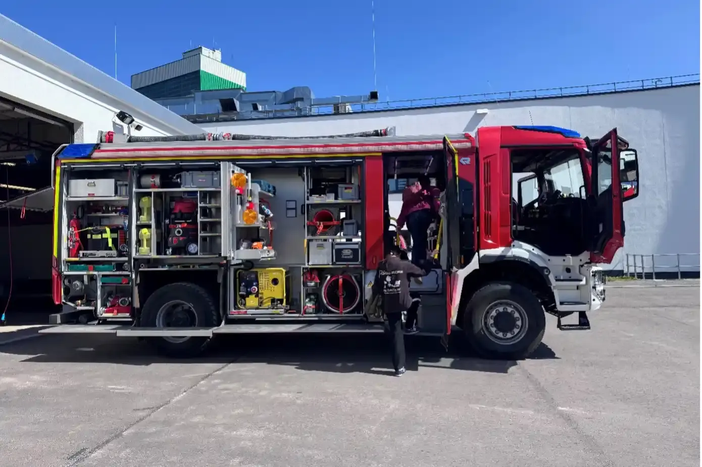 Ein Feuerwehrfahrzeug mit geöffneten Seitenrollos steht auf einem Industriegelände vor einem blauen Himmel.