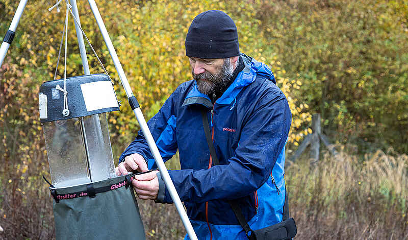 Ein Mann mit blauer Wetterkleidung hängt eine Insektenfalle an einem Dreibein in einer Waldumgebung auf.
