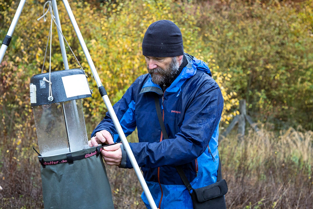 Ein Mann mit blauer Wetterkleidung hängt eine Insektenfalle an einem Dreibein in einer Waldumgebung auf.