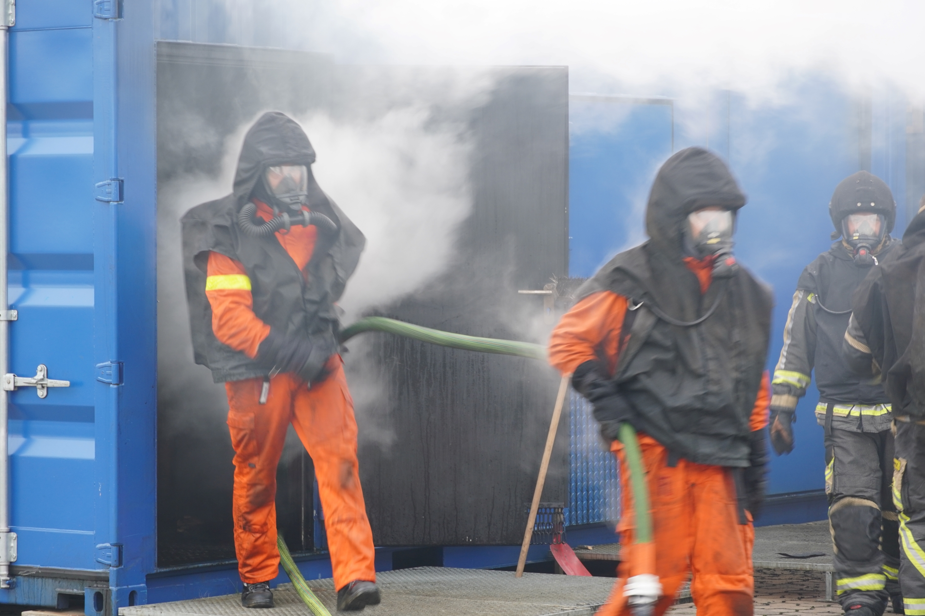 Aus einem rauchenden Container kommen mehrere Personen in Schutzkleidung und mit Atemschutzmasken.
