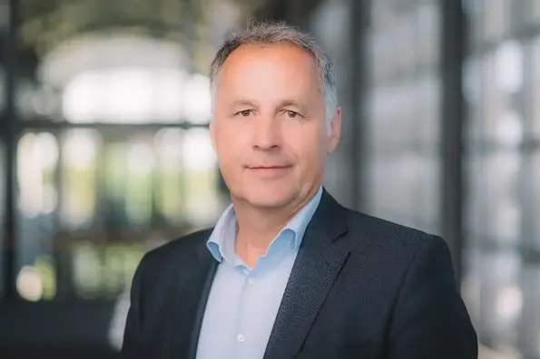 Porträtfoto eines Mannes mit kurzen grauen Haaren. Er trägt ein schwarzes Sakko und darunter ein hellblaues Hemd. 