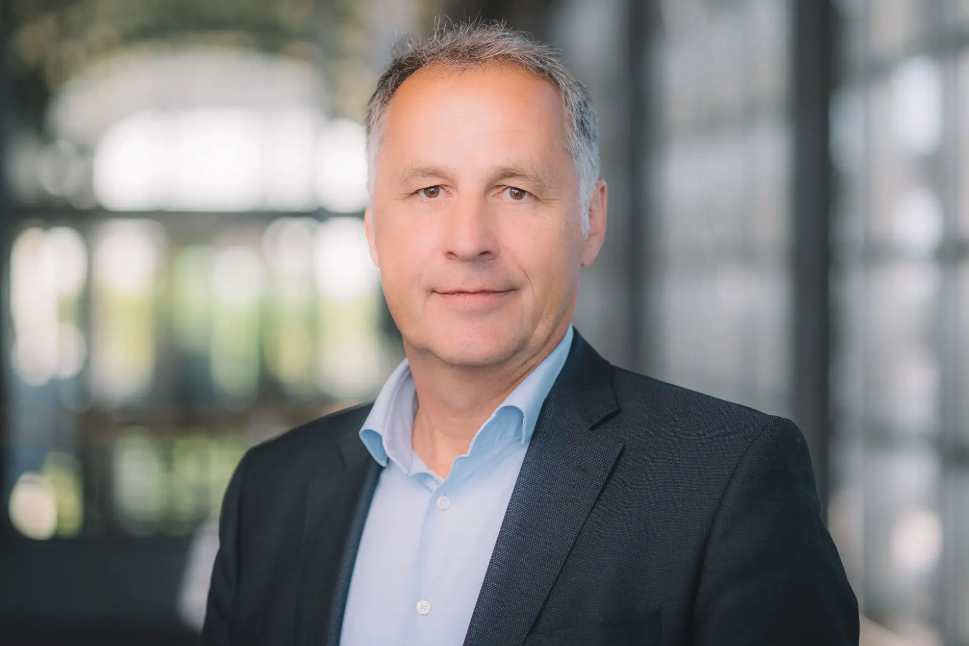Porträtfoto eines Mannes mit kurzen grauen Haaren. Er trägt ein schwarzes Sakko und darunter ein hellblaues Hemd. 