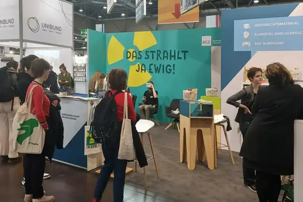 Zu sehen ist der Messestand der BGE auf der Leipziger Buchmesse 2025. Im Hintergrund ist eine Person mit VR-Brile zu sehen, rechts ein Gespräch zwischen Besucherin und einer BGE-Mitarbeiterin und im Vordergrund zwei Besucherinnen, die auf den Stand schauen.