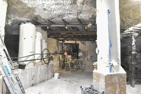 Das Foto zeigt den derzeitigen Arbeitsbereich. Im Vordergrund und auf der rechten Seite befinden sich Stützbauwerke aus Salzbeton. Im hinteren Bereich ist die eigentliche Lösungsfassung. An der Wand aus Salz ist eine schwarze Plane befestigt, um die Fassungsstelle zu schützen. Davor ist ein kleines Stehpodest aus Metall aufgestellt. Die gefasste Lösung wird über eine schwarze Rohrleitung geleitet. Das Rohr erinnert vom Durchmesser an einen größeren Gartenschlauch.