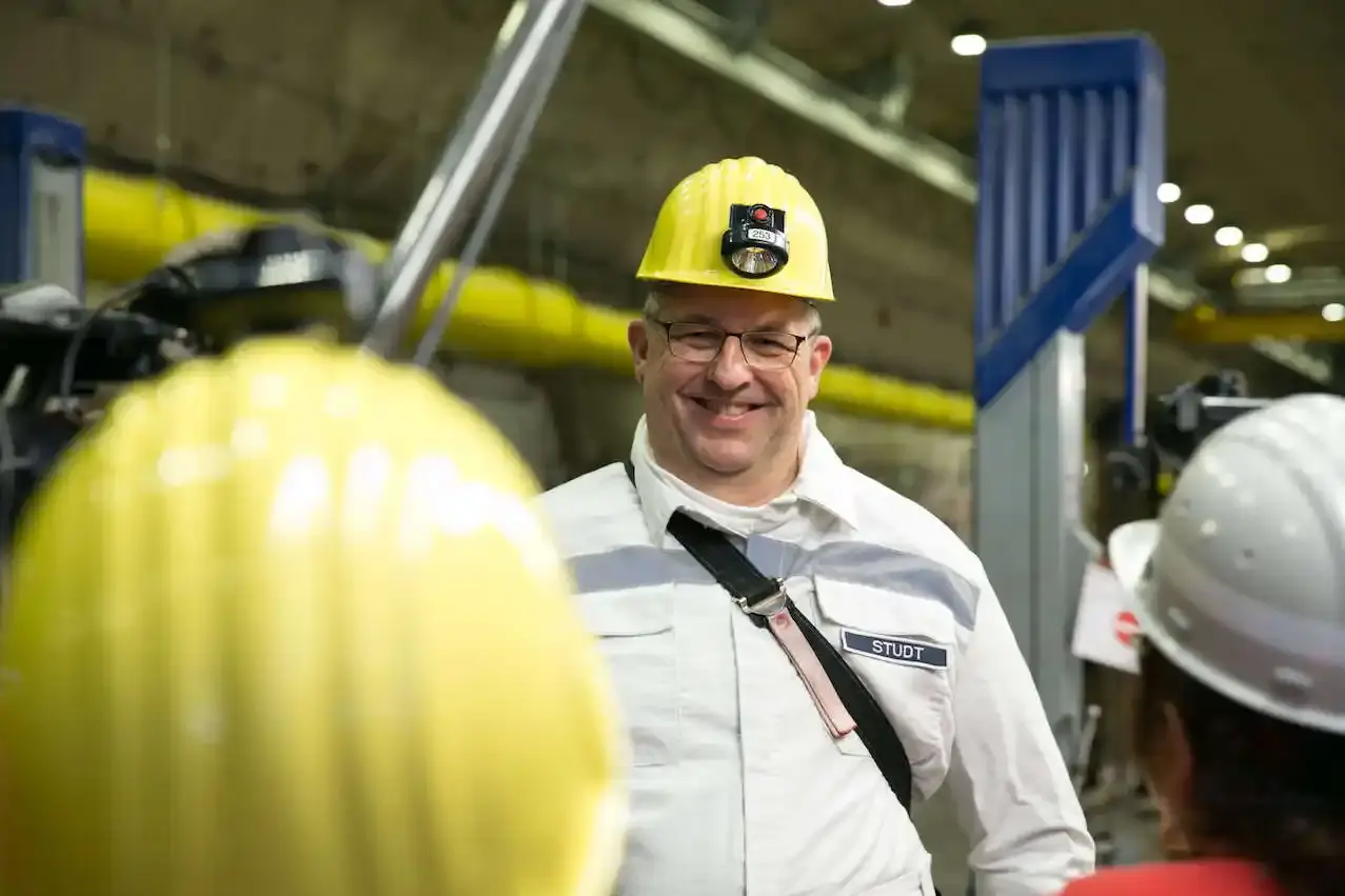 Mann in weißer Schutzkleidung mit gelbem Helm und Namensschild 'Studt' lächelt in einem unterirdischen Arbeitsbereich. Von hinten sind die Köpfe von zwei Personen mit Schutzhelmen zu sehen, die dem Mann zugewandt sind.