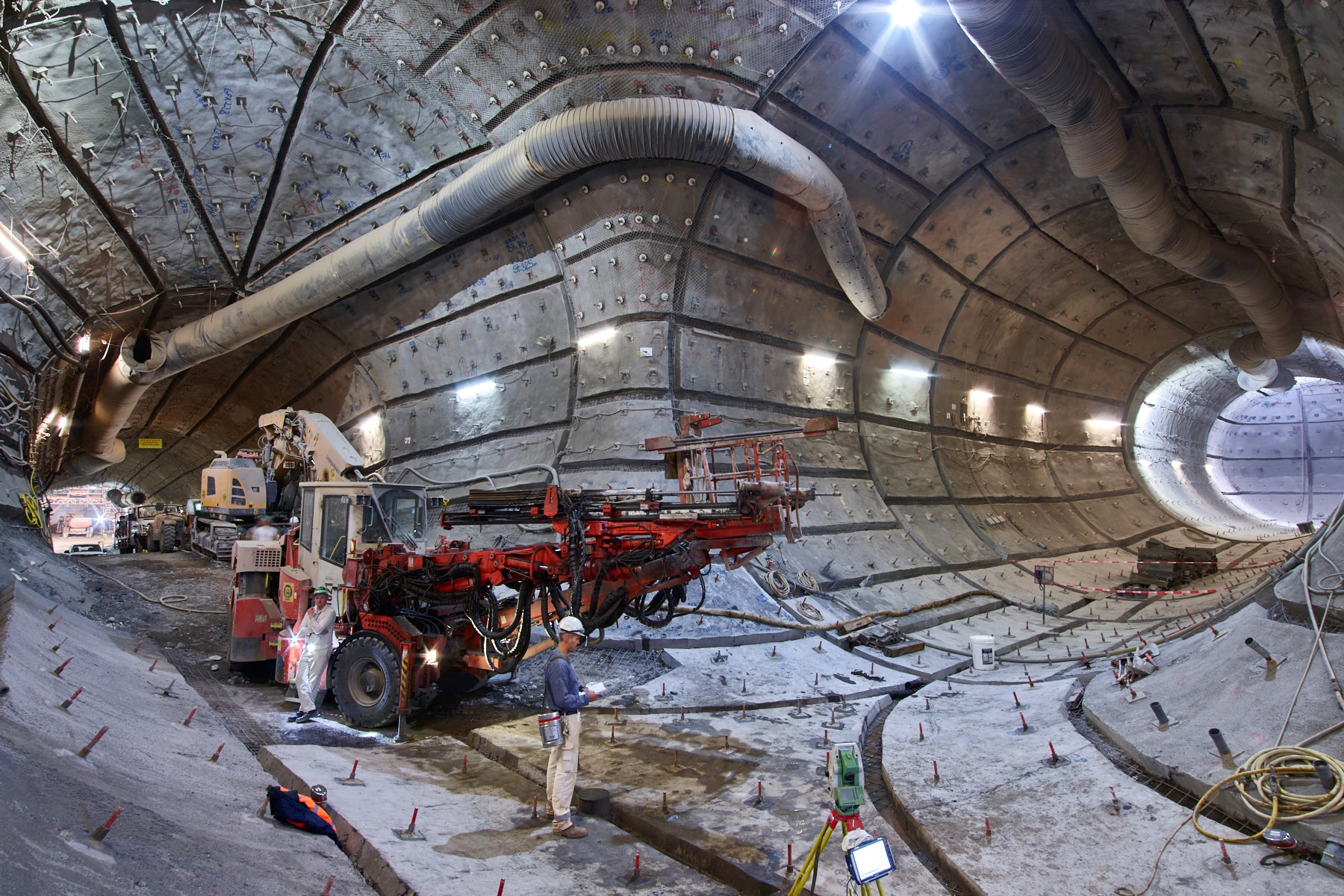 Large machines are parked in a developed section of the mine. Countless rock bolts protrude from the walls. Two miners in work clothes stand in the foreground. 