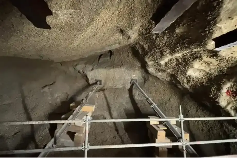 Das Bild zeigt den Blick in einen Abbau im Bergwerk. Der Boden steigt schräg in Richtung Decke an und ist mit Schotter bedeckt. Die Decke selbst besteht aus gräulichem Salzgestein. In der Bildmitte stoßen Boden und Decke aufeinander. Zentral im Übergangsbereich ragt ein Steinblock heraus. Dieser wird von zwei schräg vom Boden ausgehenden Stützen aus Metall gesichert. Im Vordergrund sind Teile eines Gerüstes und am oberen Bildrand weitere Stahlträger zu erkennen.