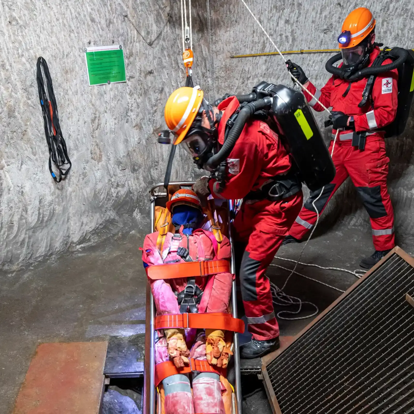 Festgeschnallt auf einer Bahre liegt eine Puppe. Zwei Personen mit Helm und Atemschutz seilen sie ab.