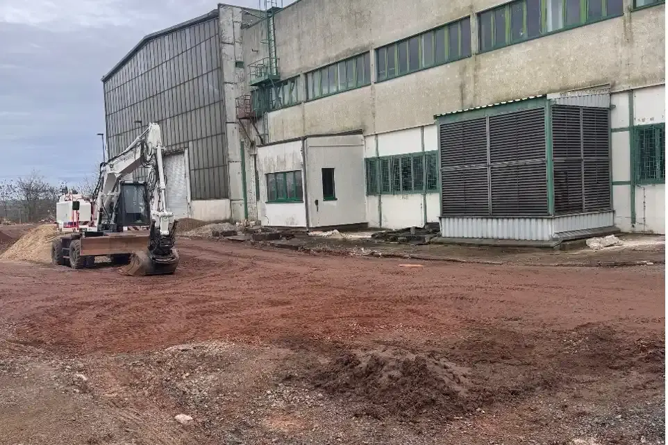 Das Bild zeigt die Außenaufnahme eines Industriegebäudes. Vor dem Gebäude steht ein weißer Radbagger auf einem verdichteten Grund. Der Himmel ist bedeckt.