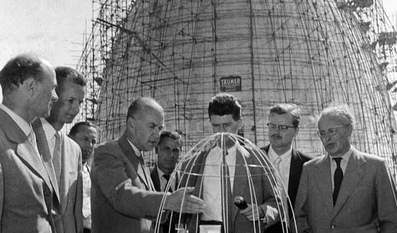 Auf dem Schwarz-Weiß-Foto stehen mehrere Männer im Halbkreis um ein Modell des Atomreaktors. Im Hintergrund ist dieser eiförmige Atomreaktor mit einem Baugerüst umgeben zu sehen.