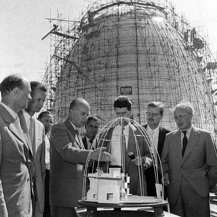 Auf dem Schwarz-Weiß-Foto stehen mehrere Männer im Halbkreis um ein Modell des Atomreaktors. Im Hintergrund ist dieser eiförmige Atomreaktor mit einem Baugerüst umgeben zu sehen.