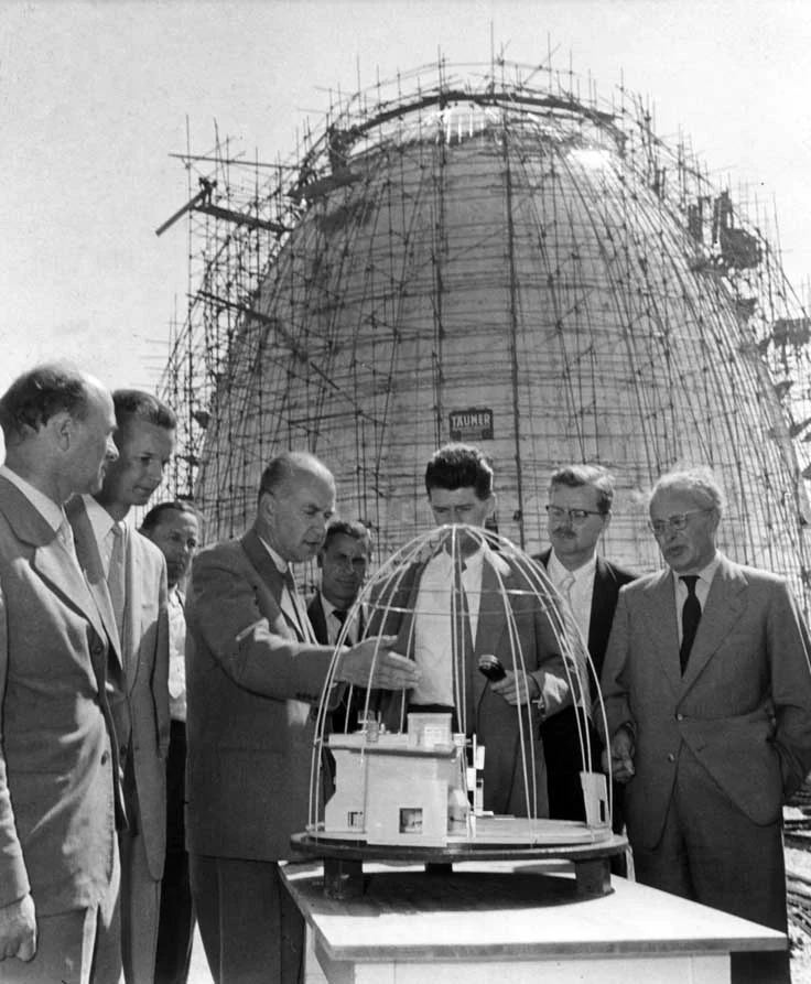 Auf dem Schwarz-Weiß-Foto stehen mehrere Männer im Halbkreis um ein Modell des Atomreaktors. Im Hintergrund ist dieser eiförmige Atomreaktor mit einem Baugerüst umgeben zu sehen.