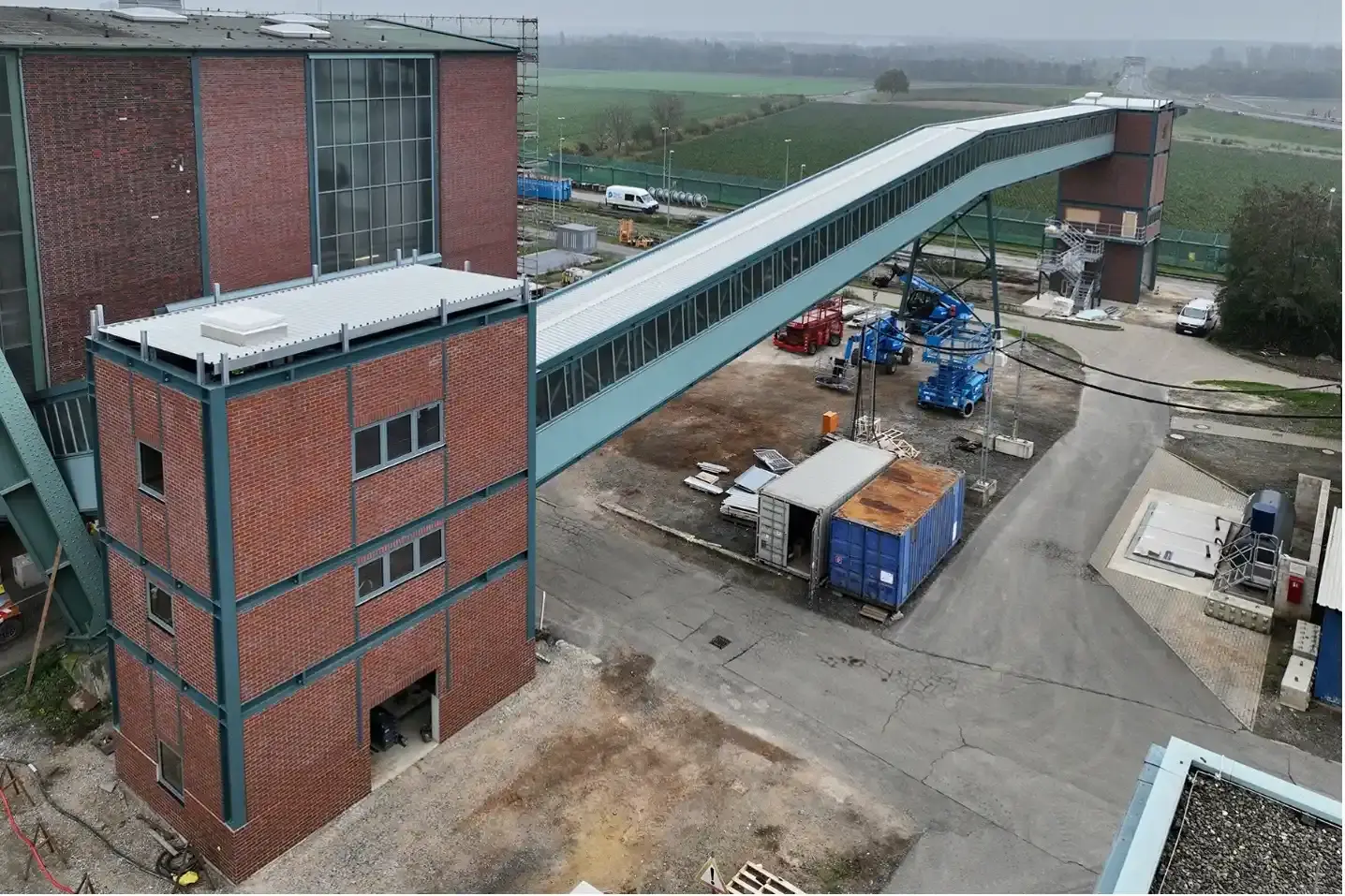 Auf einem Betriebsgelände befinden sich rote Backsteingebäude und eine Bandbrücke, die zwei dieser Gebäude miteinander verbindet.