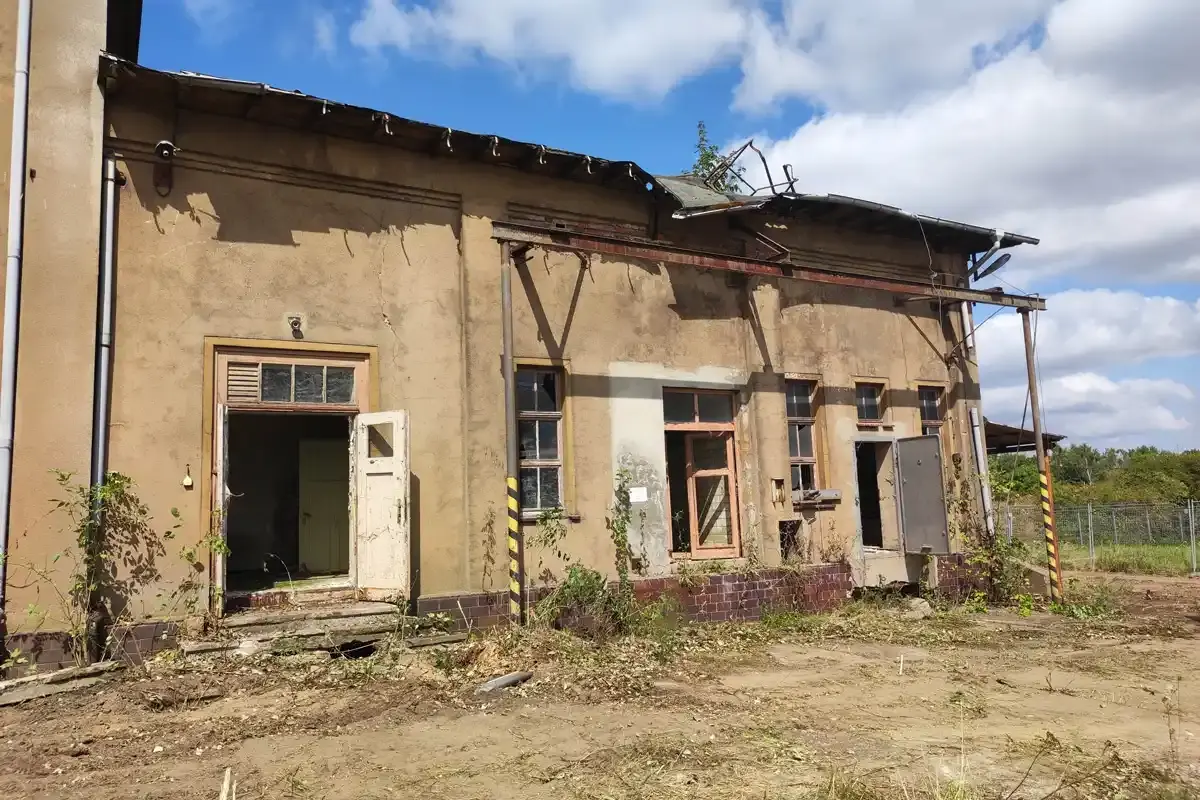 Frontalansicht eines alten Gebäudes mit starkem Verfall. Von den geöffneten Eingangstüren blättert die weiße Farbe ab.