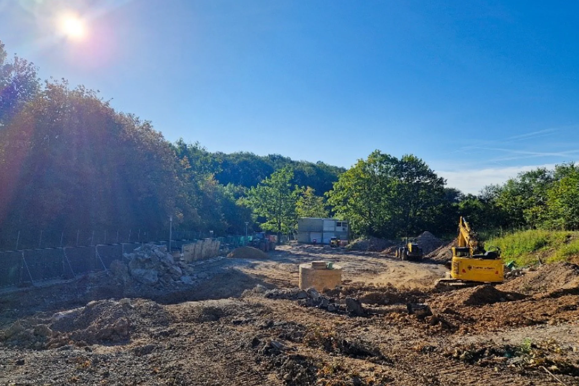 Das Bild zeigt eine Freifläche mit aufgegrabener Erde, eingerahmt von grünen Bäumen. Innerhalb der Freifläche stehen Baufahrzeuge. Am linken Bildrand liegt ein Haufwerk aus Asphaltresten. Im hinteren Bereich steht ein zweistöckiger Baucontainer.   