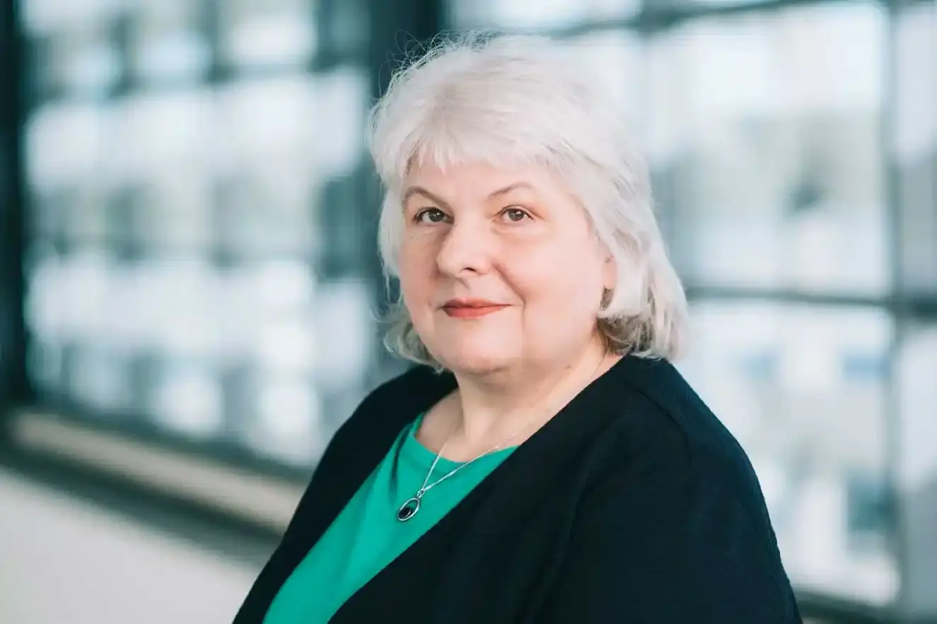 Eine Frau mit grauen schulterlangen Haaren steht vor einer großen Fensterfront und blickt freundlich in die Kamera. Sie trägt ein grünes Oberteil unter einer schwarzen Strickjacke. 