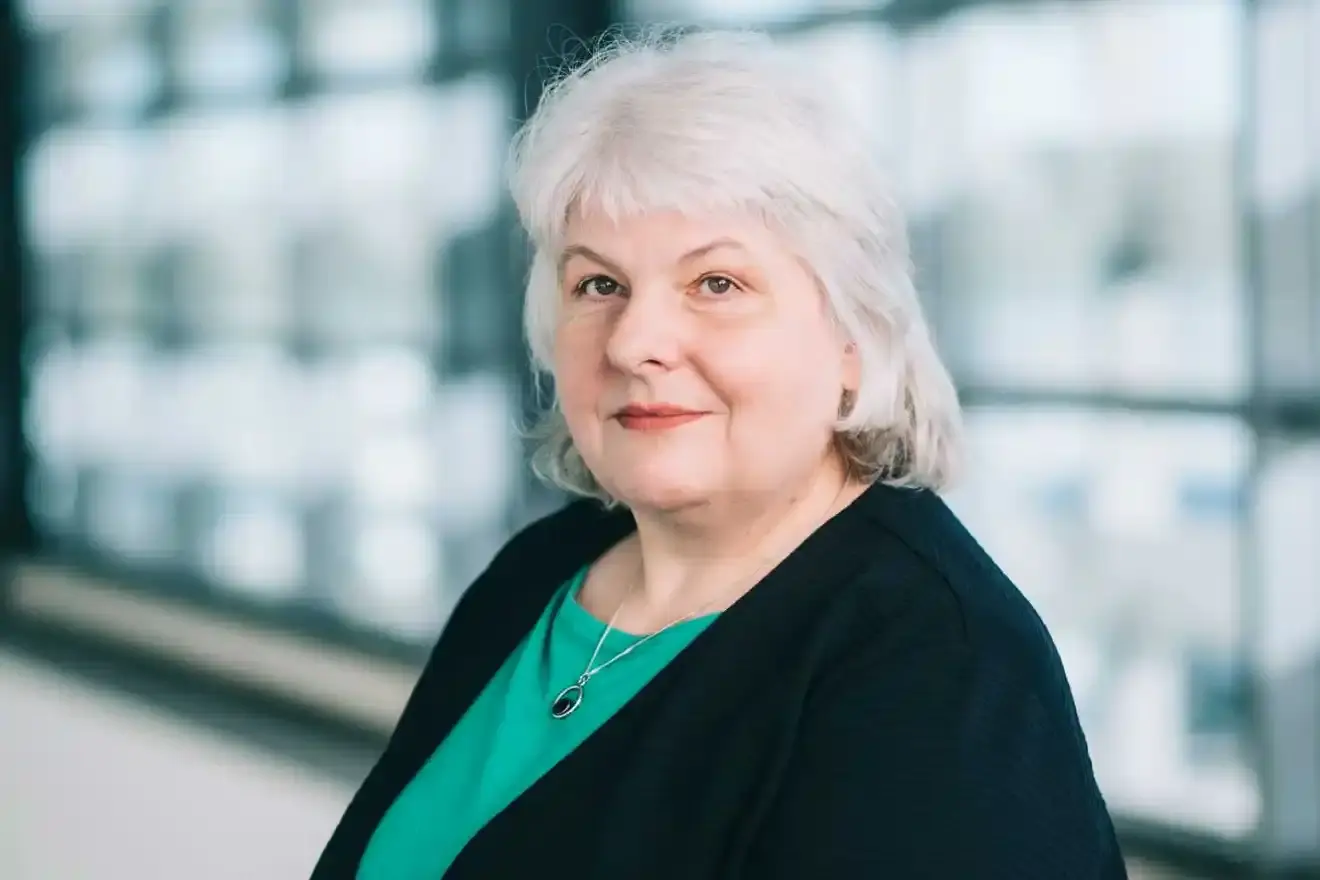 Eine Frau mit grauen schulterlangen Haaren steht vor einer großen Fensterfront und blickt freundlich in die Kamera. Sie trägt ein grünes Oberteil unter einer schwarzen Strickjacke. 