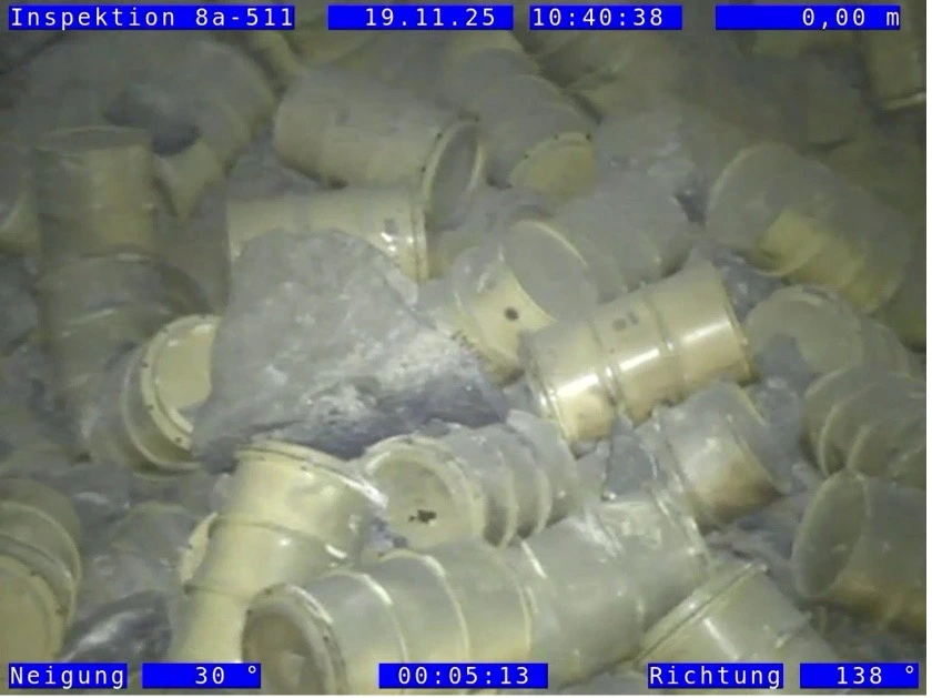 Das Bild zeigt durcheinanderliegende gelbe Fässer in einer Einlagerungskammer. Die Fässer sind zum Teil mit größeren und kleineren Brocken aus Salzgestein und Staub bedeckt. Im oberen und unteren Bereich des Bildes befinden sich Display-Einblendungen mit technischen Informationen auf blauem Grund.