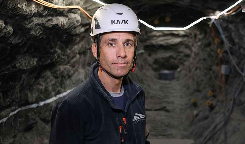 Mann mit weißem Helm und schwarzer Jacke blickt unter Tage in die Kamera. Im Hintergrund sind Gesteinswände mit Kabeln zu sehen.