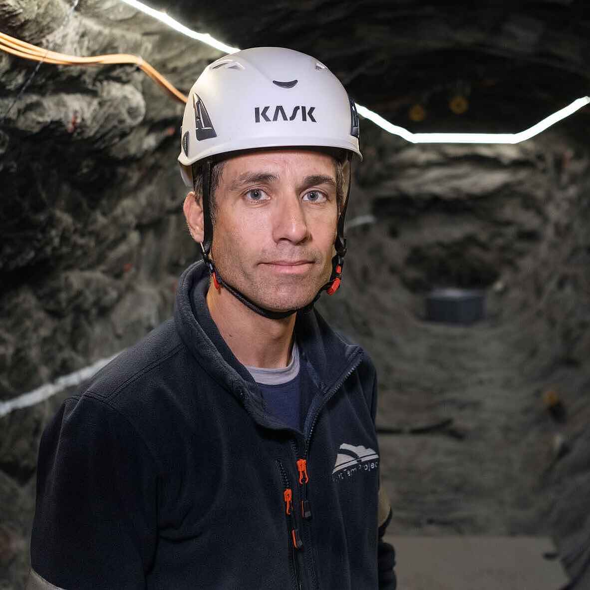 Mann mit weißem Helm und schwarzer Jacke blickt unter Tage in die Kamera. Im Hintergrund sind Gesteinswände mit Kabeln zu sehen.