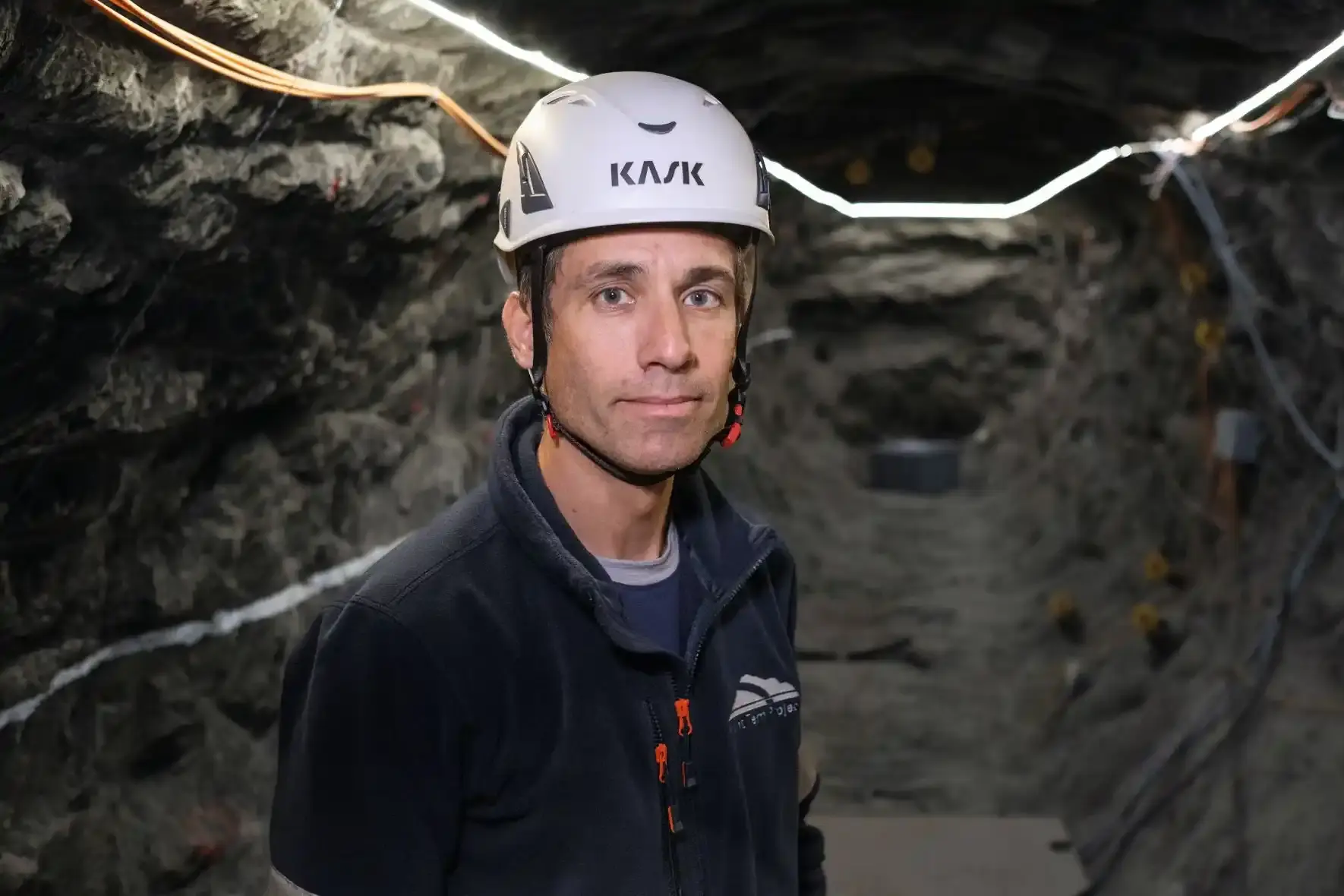 Mann mit weißem Helm und schwarzer Jacke blickt unter Tage in die Kamera. Im Hintergrund sind Gesteinswände mit Kabeln zu sehen.