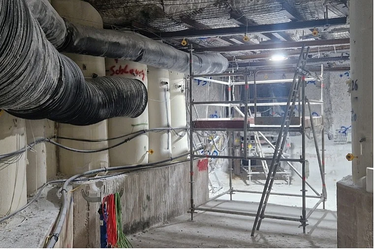 Das Bild zeigt einen Arbeitsbereich in einem Bergwerk. Auf der linken und rechten Seite befinden sich pfeilerartige Stützbauwerke aus weißgrauem Salzbeton. Ein Metallgerüst steht in der Mitte des Fotos. An den Wänden und der Decke verlaufen Schläuche und Balken.