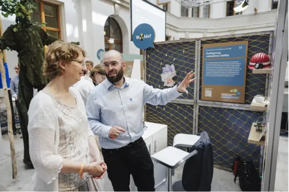 Eine Informationswand mit Texten und Exponaten zur BGE. Vor der Informationswand steht ein BGE Mitarbeiter und erläutert einer Besucherin die Inhalte.