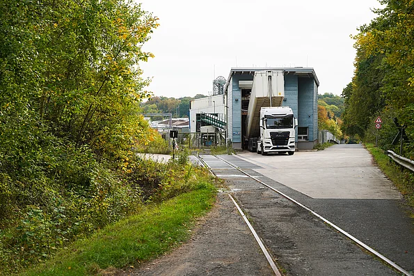 Lkw vor einem Industriegebäude an einer Straße mit Gleisen, umgeben von Bäumen.