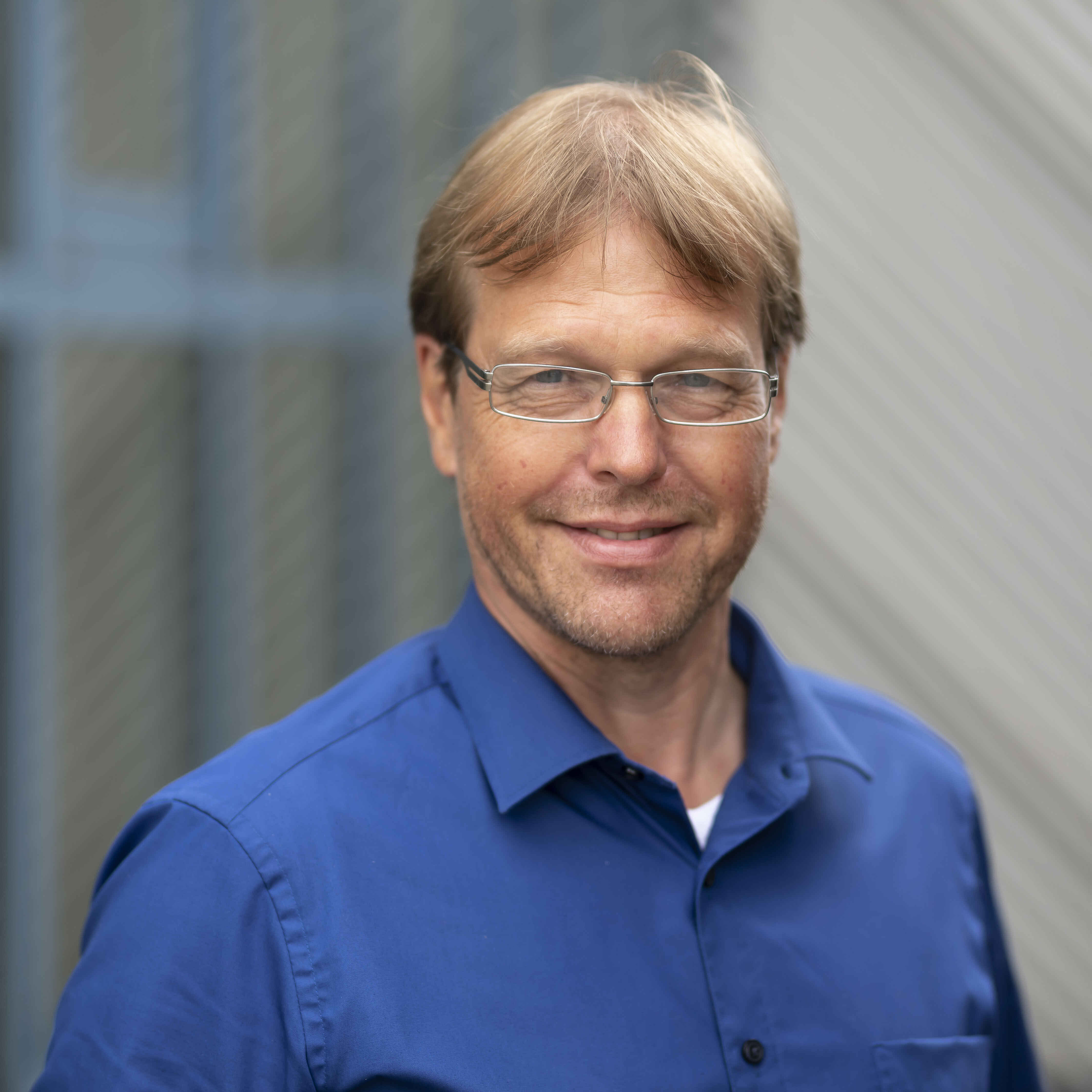 Portraitfoto von einem Mann mit Brille, dunkelblondem Haar und blauem Hemd.