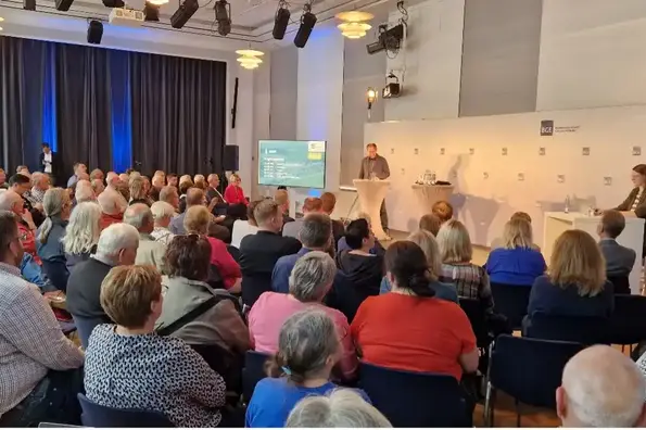 Eine Gruppe von Menschen sitzt in einem Saal und schauen auf eine Bühne. Vorne stehen mehrere Personen an Rednerpulten.