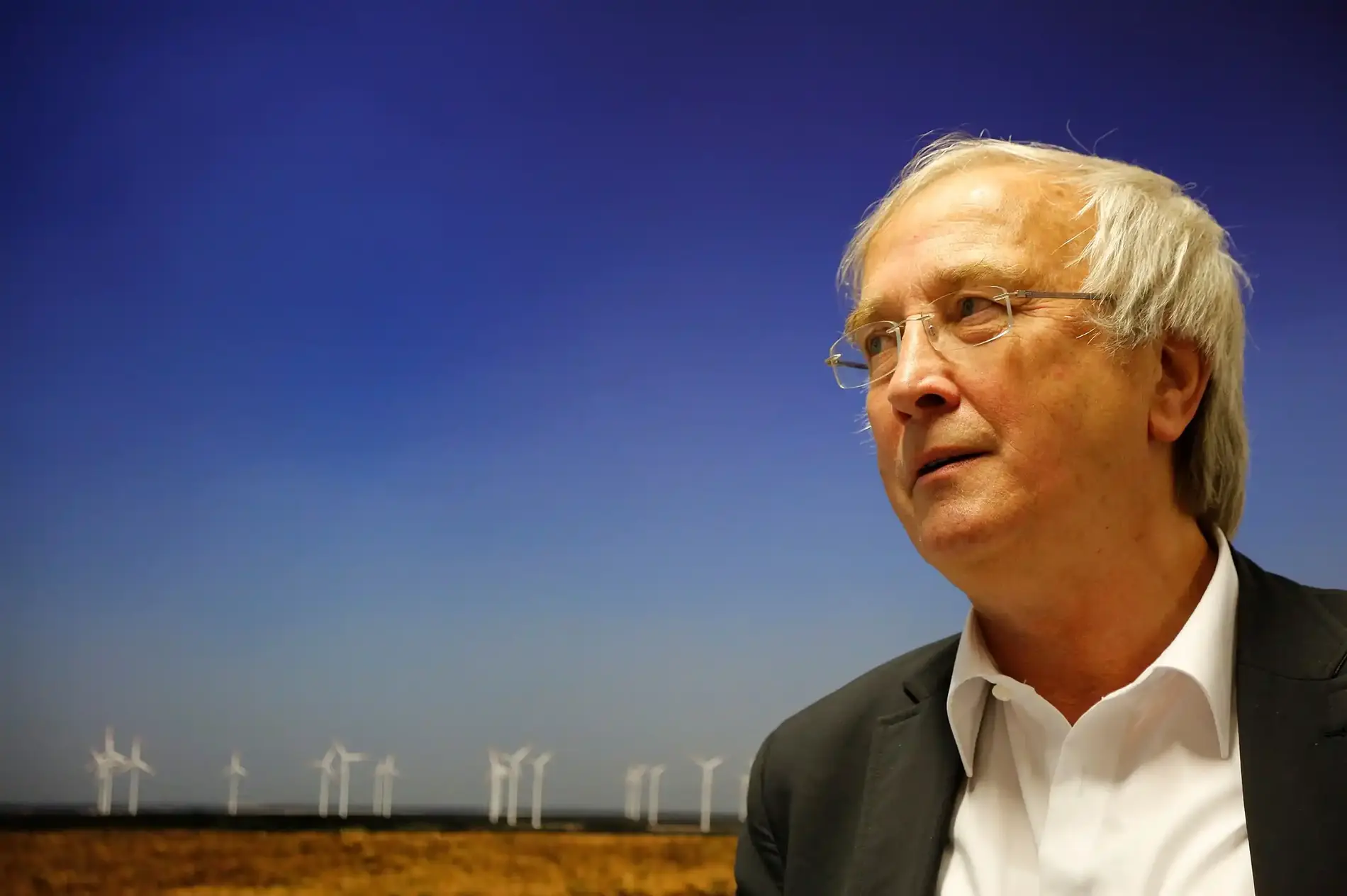Portraitbild von Raimund Kamm: Mann mit weißen Haaren und Hemd vor einem blauen Hintergrund mit Windrädern.