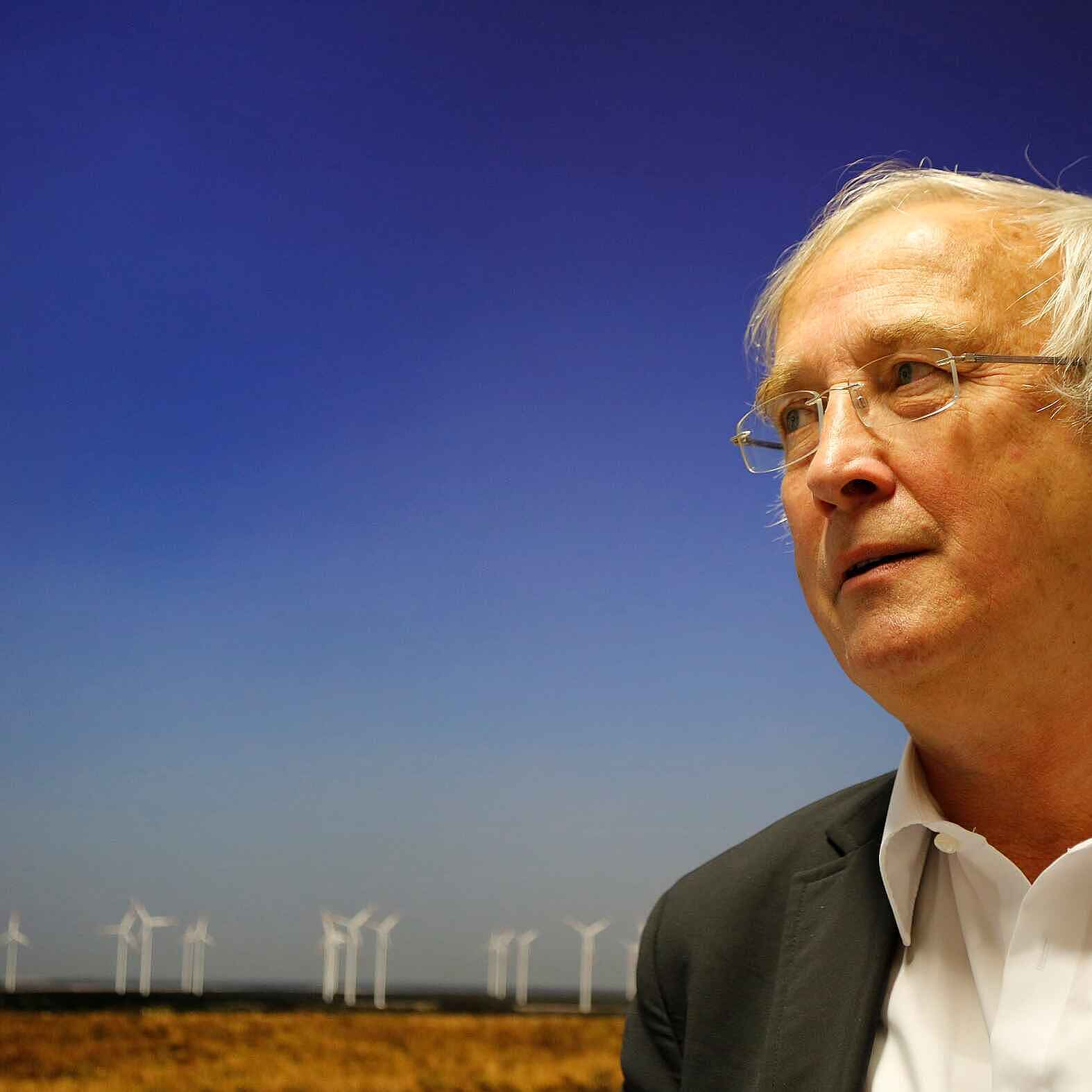 Portraitbild von Raimund Kamm: Mann mit weißen Haaren und Hemd vor einem blauen Hintergrund mit Windrädern.