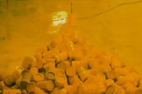 Das Bild zeigt gelbe Fässer, die in einem kegelförmigen Haufen in einer Kammer liegen. Ein Fass wird oberhalb des Fasskegels an einem Stahlseil herabgelassen.