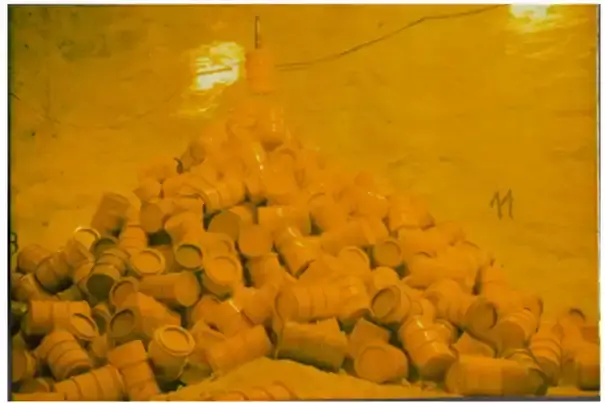 Das Bild zeigt gelbe Fässer, die in einem kegelförmigen Haufen in einer Kammer liegen. Ein Fass wird oberhalb des Fasskegels an einem Stahlseil herabgelassen.