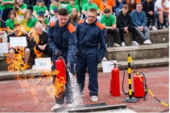 Zwei junge Männer in Schutzanzügen und Schutzbrillen löschen mit einem Feuerlöscher ein offenes Feuer auf einem Übungsplatz. Im Hintergrund sitzen Zuschauer*innen auf Betonstufen.