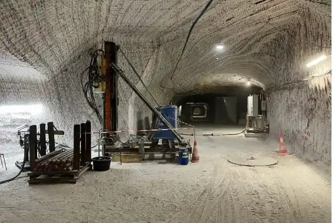 Das Bild zeigt einen Tunnel, der tief in das Bergwerk führt. Im vorderen, linken Bereich ist eine Nische in das Salz gefräst. Darin steht eine Bohranlage aus Metall. Vor der Bohranlage liegen rund ein Dutzend einzelne Bohrgestänge aus Metall in einer Transportbox. Der Boden um die Bohranlage ist mit weißem Salzstaub bedeckt. Die Wände und die Decke der Strecke zeigen typische Frässpuren und Salzstrukturen.