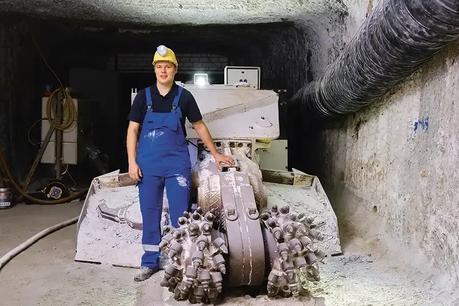 Ein junger Bergmann steht im Schacht vor einer Maschine. Erträgt blaue Latzhose und einen gelben Helm mit Stirnlampe.