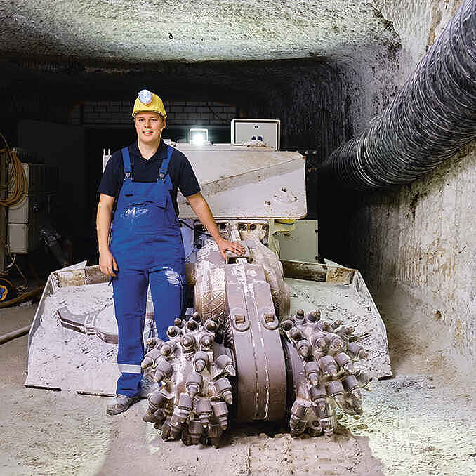 Ein junger Bergmann steht im Schacht vor einer Maschine. Erträgt blaue Latzhose und einen gelben Helm mit Stirnlampe.