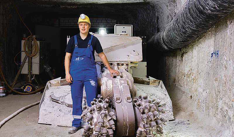 Ein junger Bergmann steht im Schacht vor einer Maschine. Erträgt blaue Latzhose und einen gelben Helm mit Stirnlampe.