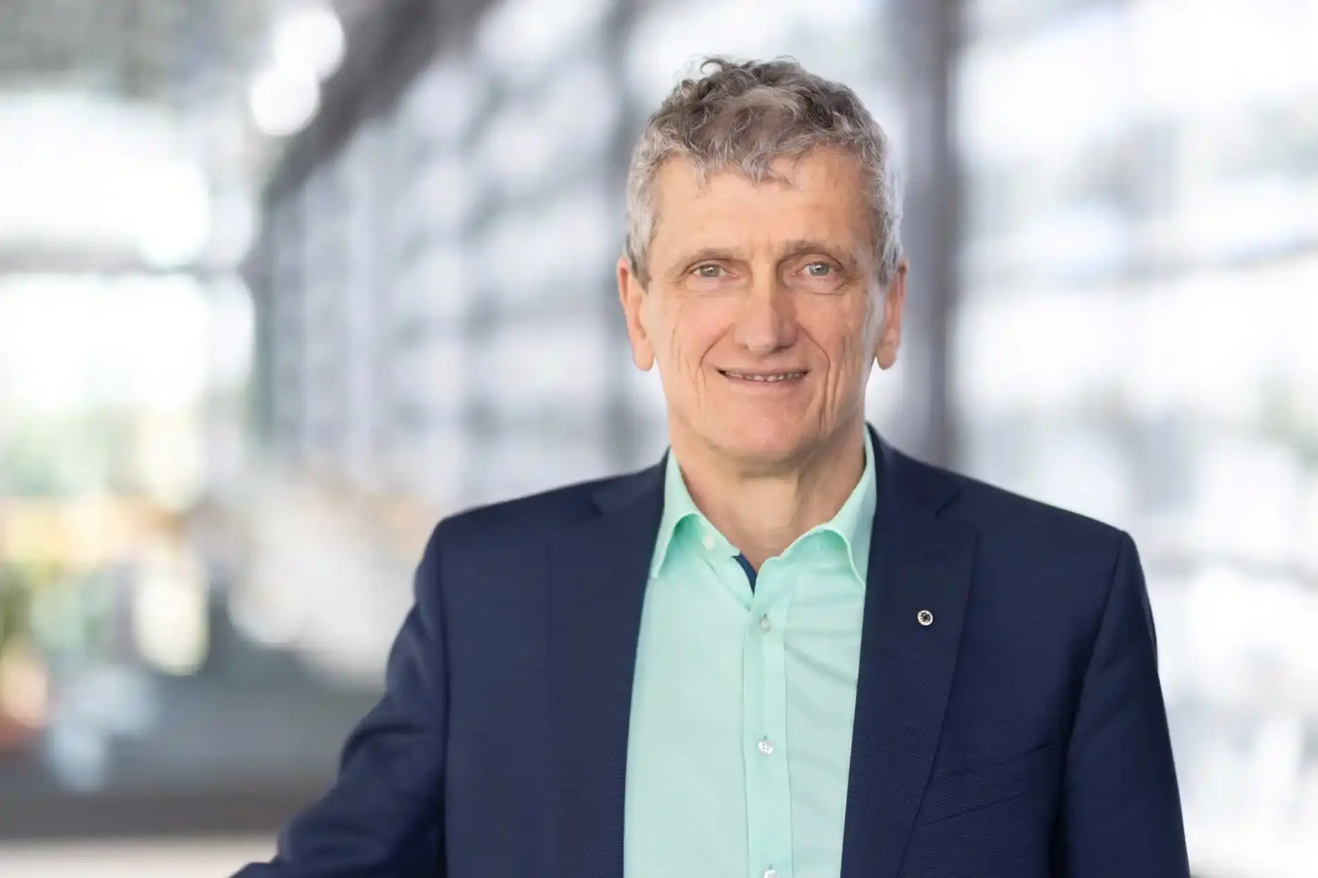 Portrait photo: A man with short gray hair, a green shirt and a dark blue jacket smiles at the camera.